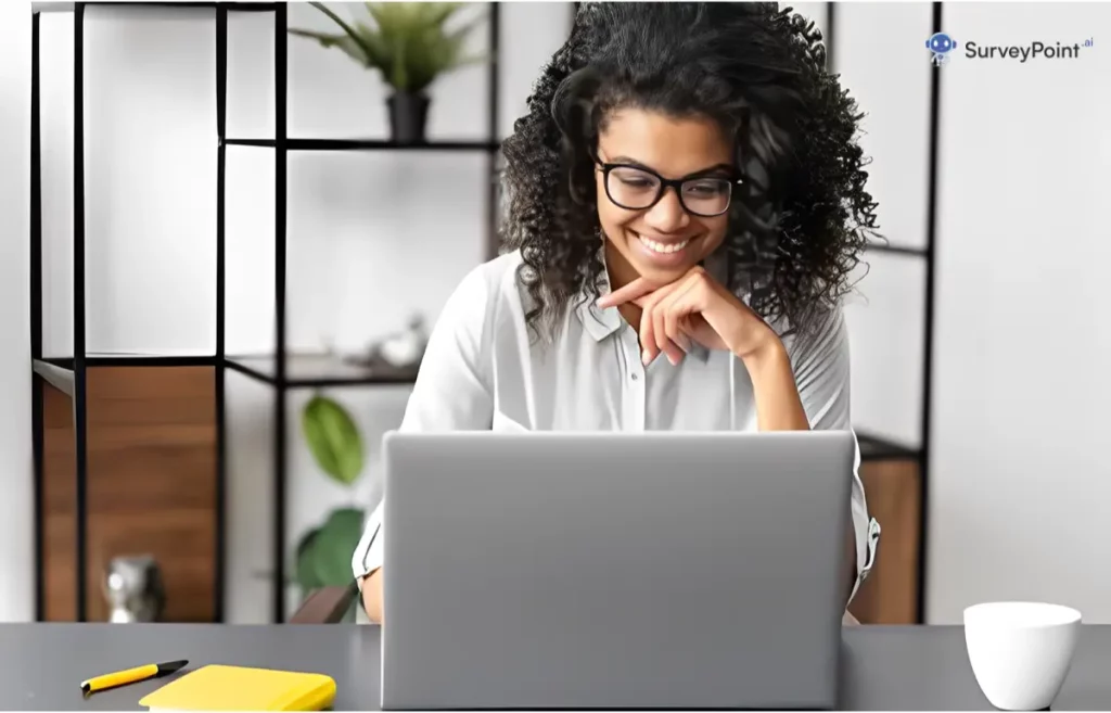 A woman with curly hair sits at a desk, focused on her laptop, surrounded by materials for pre and post surveys.