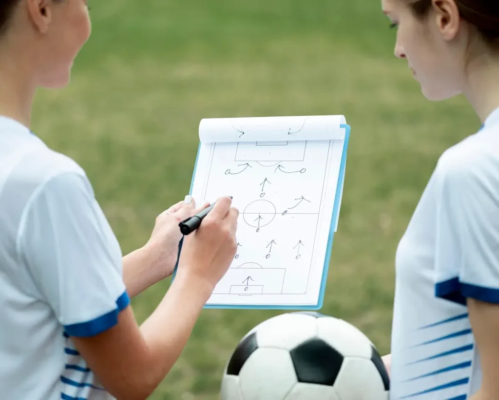 Two female soccer players review a clipboard, discussing strategies from Crack the Performance Code sports survey questions.